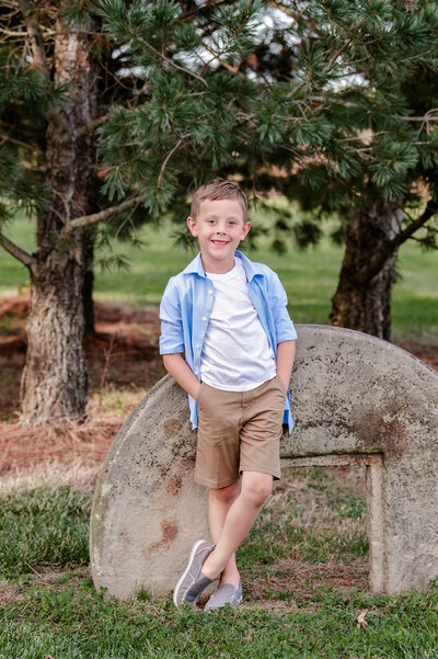 Little boy leans against concrete statue with ankles crossed smiling with hands in his pockets.