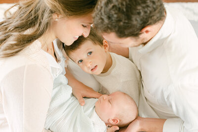 Portrait of a man and a woman wearing all white smiling down at a boy and a swaddled newborn baby nestled in between them