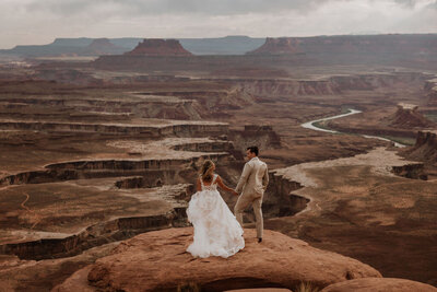 bride and groom standing on rock formation on the side of a cliff facing the desert
