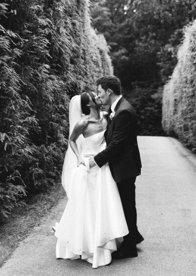 black and white photo of bride and groom kissing in between hedges 