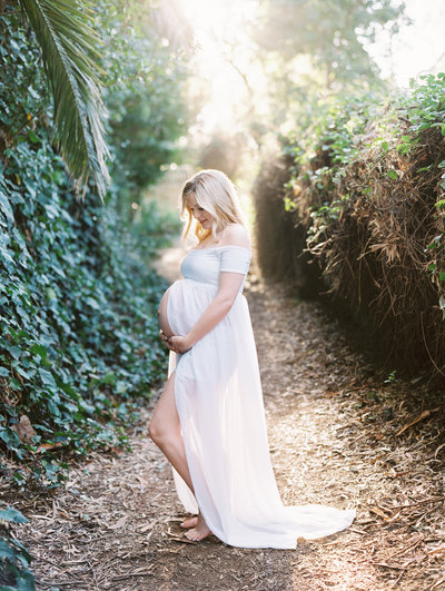 A mom in a white dress smiles at her growing belly during her maternity session with Daniele Rose