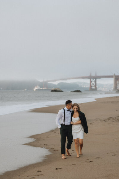 A couple walks barefoot on a foggy beach during their elopement photoshoot, the man in a white shirt and suspenders, the woman in a short white dress and black shawl, with the Golden Gate Bridge in the background