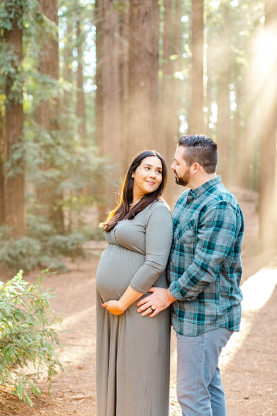 Pregnant woman holds baby shoes in front of her stomach while her partner embraces here from behind.