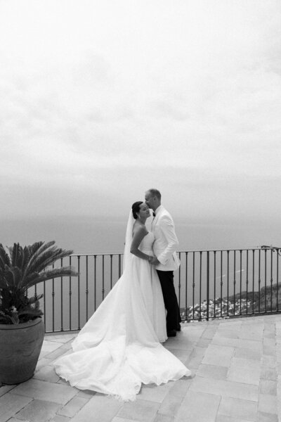 Bride and groom kissing on a balcony overlooking the city of Positano, Italy on their wedding day, captured by Andrea-Marie Photography