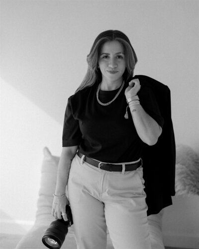 Black and white portrait of a destination elopement photographer confidently holding a camera, dressed in neutral tones with a blazer draped over her shoulder, standing in a softly lit modern space.