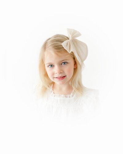 A sweet blonde girl wearing a soft ivory bow gazes forward in a bright and airy portrait setting.
