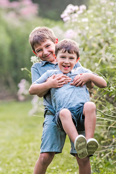 Older brother picks up little brother while both are smiling and laughing surrounded by spring foliage
