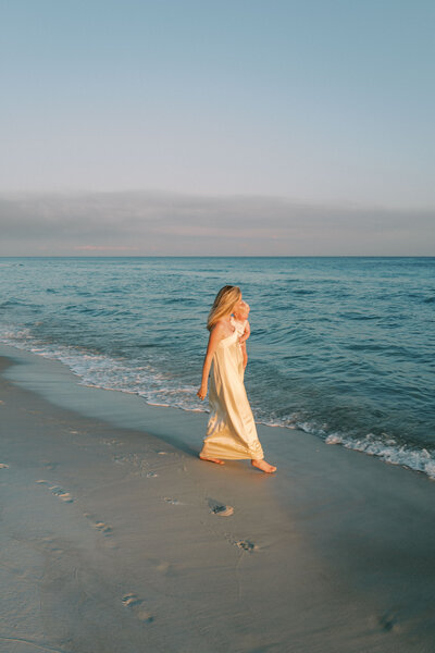 Mom in long dress holding baby and walking in the sand beside the ocean