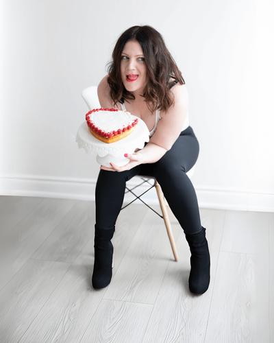 Emily licks crumbs off her upper lip while sitting on a chair with a heart-shaped cake.