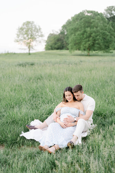 pregnant mother cuddles husband in a field in a maternity session with AnneMarie Hamant
