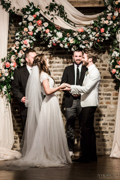 Bride and groom hold hands and laugh during wedding ceremony under lush pink and white draped floral archway