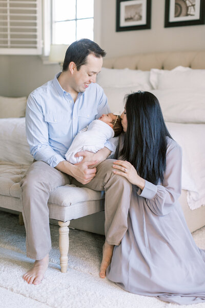 A father sits on a bench holding his newborn daughter while the mother kneels beside them and kisses the baby's head during their newborn session