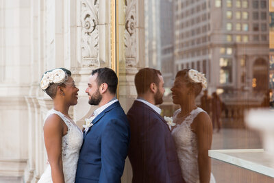 Groom leaning on a wall glass while bride is leaning on him smiling at each other