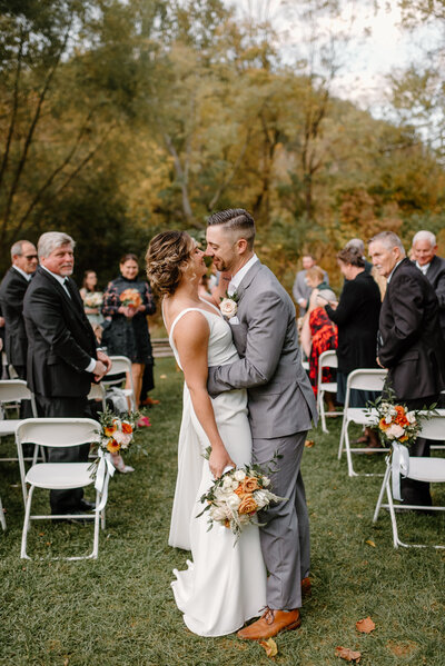 Bride and groom going in for a kiss at the end of their recessional at the Philander Chase Knox Estate wedding in Valley Forge, Pa. The groom has his arms wrapped around the brides waist and smiling at her as their noses touch. The bride has her left arm up on his face and her  right arm is down holding a beautiful bouquet of bridal flowers with oranges and white florals. The groom is wearing a gray suit with  light brown leather shows . The bride is wearing a form fitting white stain dress with a cape and her hair is in a flowy updo. Greater philadelphia wedding ceremony and wedding venue. Wedding photographs of a couple just married.