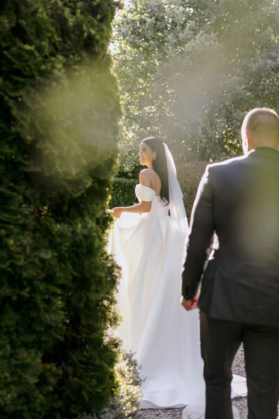Bride in beautiful light wearing Trish Peng gown disappearing behind the hedge followed by her groom