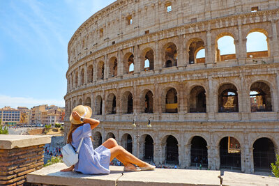 A person in a blue dress and hat sits on a ledge, looking at the Colosseum in Rome. The ancient structure's arches and stonework are visible against a clear blue sky.