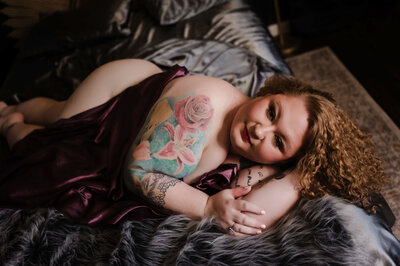 A woman with tattoos snuggles a purple sheet while laying across a bed under a window