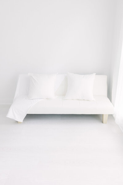 A peek inside the Looking Up Photography studio, located in Greenwich, Connecticut. A large, nearly floor-to-ceiling window bathes the studio in beautiful natural light. The center wall allows for several backdrop options, while a white chaise, rolling stool, and other photography props pepper the studio space.