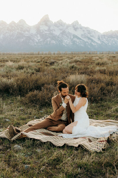 man and woman sitting on a blanket on the ground outside with mountains in the background