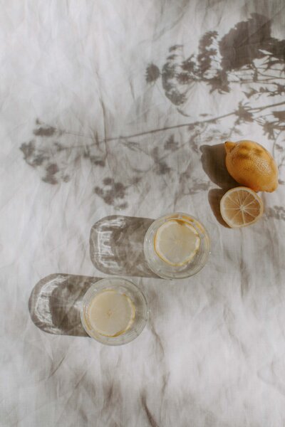 white sheet with floral shadows, glass of water with lemons, full lemon, half cut lemon