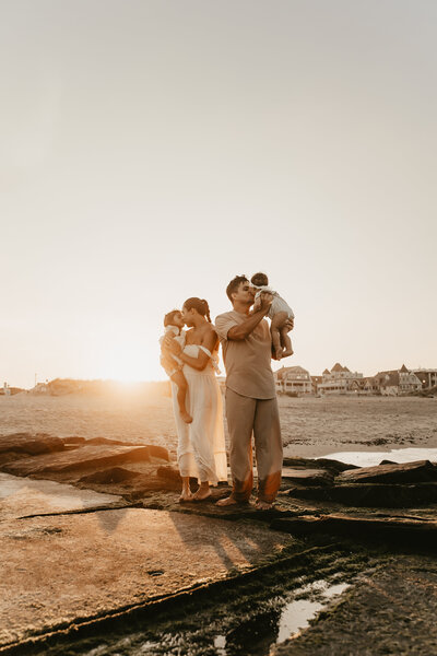 Family of five standing together on a lakeside rock during an outdoor photo session in Stanhope, NJ, with autumn trees and hills in the background, captured by Burkat Photography
