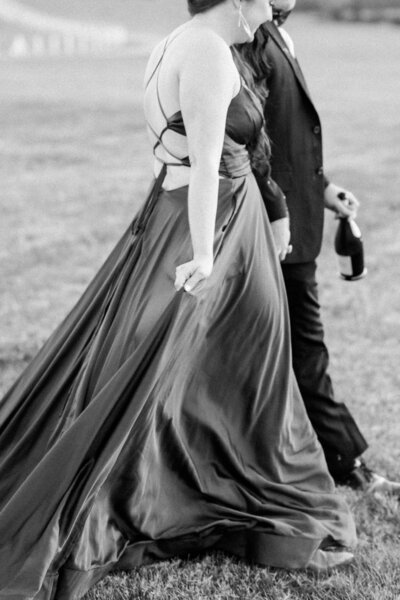  A black-and-white image of a woman in a flowing gown holding her dress while walking beside her partner, who holds a bottle of champagne