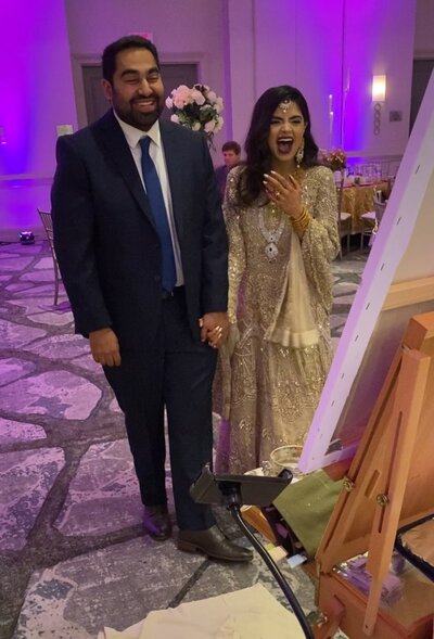 South Asian couple, bride in gold beaded gown, delightedly reacting to their live wedding painting at their reception
