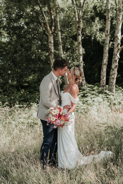 Bride & Groom standing closely facing eachother, bride holding soft summer floral bouquet in the long grass at their wedding at Oak Tree Barn