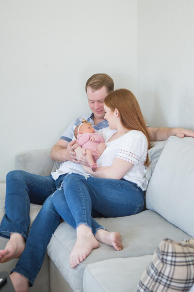 Redheaded couple lovingly snuggling newborn baby girl on gray couch at their lifestyl in home newborn session