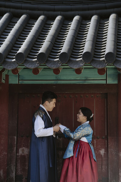 a couple in their wedding hanboks stand in front of a hanok door on their wedding day