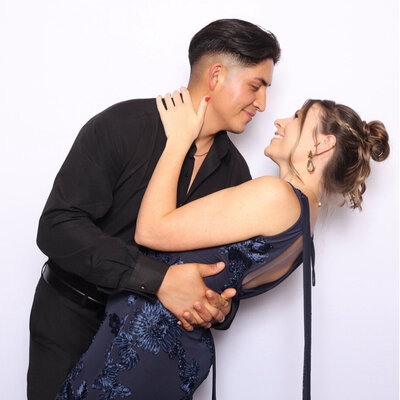 Two women in white dresses posing closely at a photo booth with a soft lavender backdrop, smiling playfully at the camera.