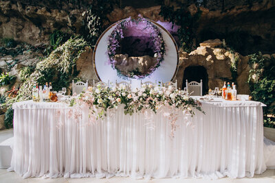 Scénographie de mariage proposée par une organisatrice d'événements. Table des mariées décorée de voilage, végétal et surmontée d'une arche en forme de croissant de lune.