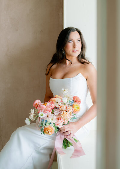 Portrait of bride sitting on a window sill holding a colorful bouquet and looking out the window