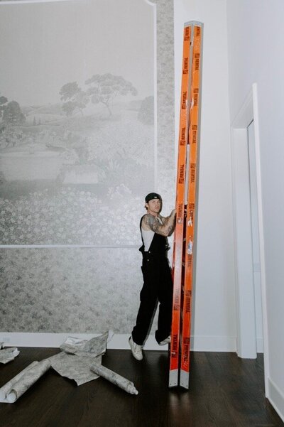 A man wearing overalls, a backwards hat posing with a ladder while hanging wallpaper