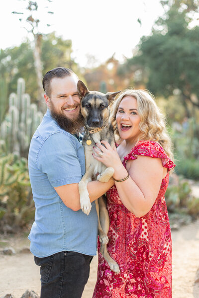 Couple holds their german shepard dog mix while smiling at the camera in a cactus garden.