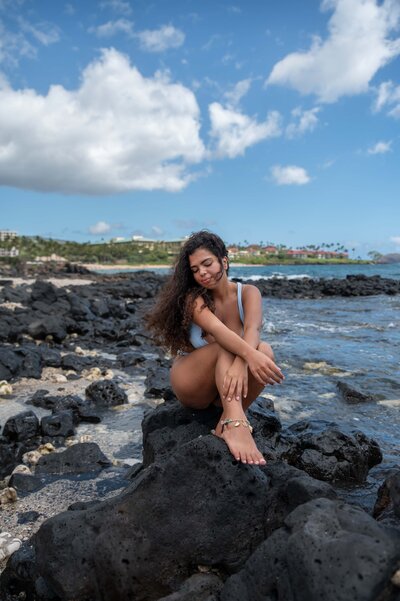 Girl in light blue bathing suit sitting on black rock looking down the side of her arm, looking pensive. 