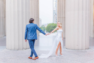 A blonde bride wearing a strapless, satin white dress that has a high slit and a big bow on the back holding hands with her fiance wearing a blue suit with brown shoes by nashville wedding photographer, brooke elliott