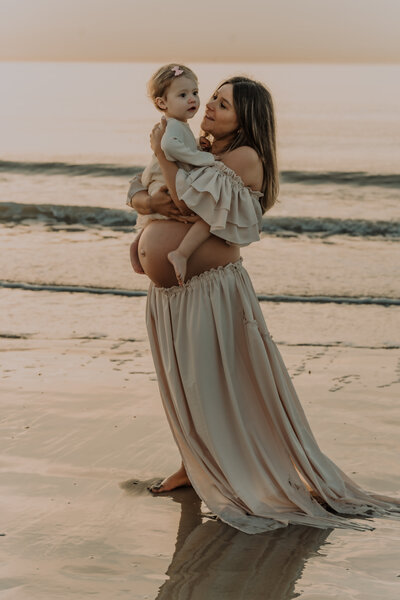 Zwangere vrouw met dochter poseert voor zwangerschapsshoot op het strand tijdens zonsondergang