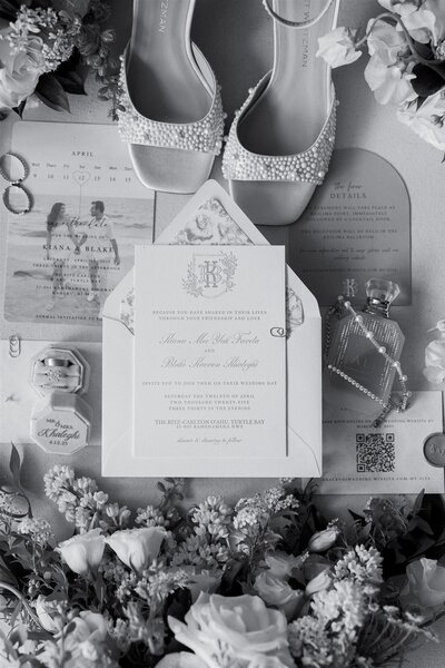 A flat lay with wedding invitations, rings, shoes, perfume, jewelry, and florals 