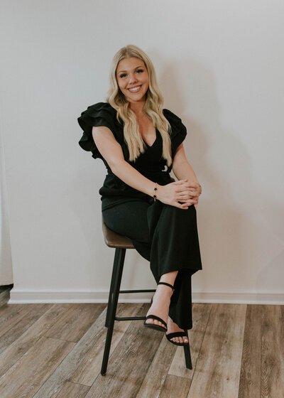 A professional headshot of Ambrin Smith, a Guest Services team member at Heath Salon and Spa. She is seated on a stool with her legs crossed, smiling warmly with a friendly and approachable demeanor. The clean background enhances her polished and professional appearance.