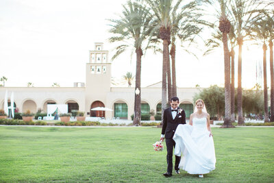 Groom holds his Bride's dress train as they walk across a large green lawn area