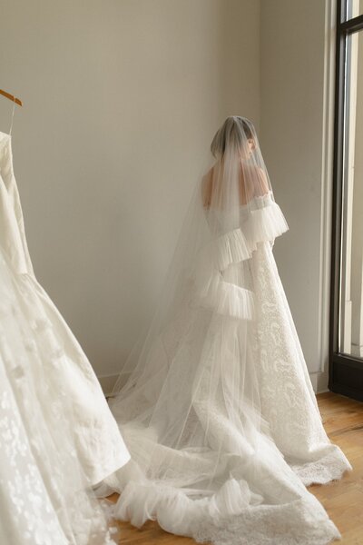 Bride in an off-shoulder lace gown with a long veil standing near a window.