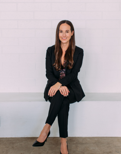 Kristie wearing all black, sitting against a white wall, smiling for the camera