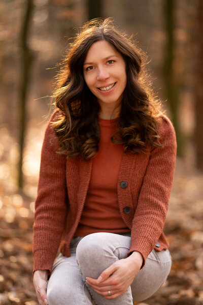 Vrouw zit op een boomstam met bladeren op de grond om haar heen en houdt haar knie vast en kijkt schuin en lachend naar de camera
