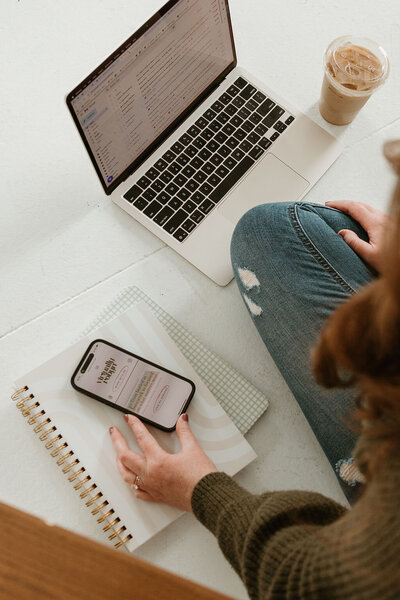 Virtually Robyn, a virtual assistant, is typing on a laptop, with an iced coffee, a smartphone, and a notebook nearby. 
