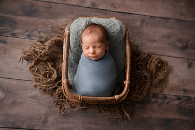 Sleeping baby boy with brown hair wrapped in a dusty blue wrap placed in wood basket