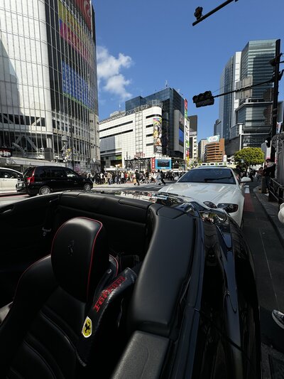 Interior view of a black Ferrari convertible in the heart of Shibuya, Tokyo, with glass towers and a bright blue sky reflecting off surrounding buildings.

