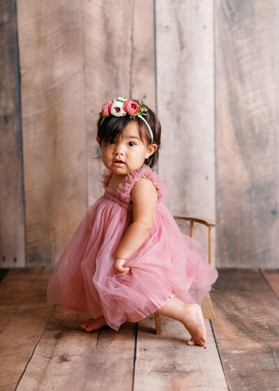 Bébé de 1 an assis sur chaise en bois et fond bois sombre, en studio. Fillette avec sa robe rose et serre tête en fleurs