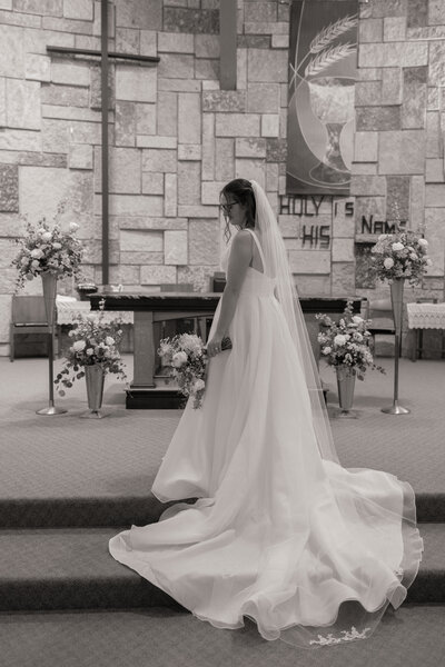 Stunning bride in a flowing white wedding gown captured at the church altar, holding a bouquet. Beautiful floral arrangements surround the bride, perfect for classic and elegant wedding photography.