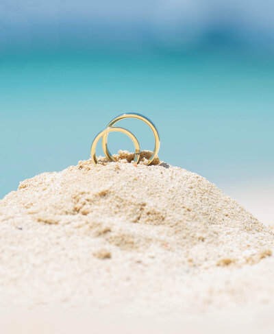 Two gold wedding rings placed on a small mound of sand with a blurred turquoise ocean in the background.